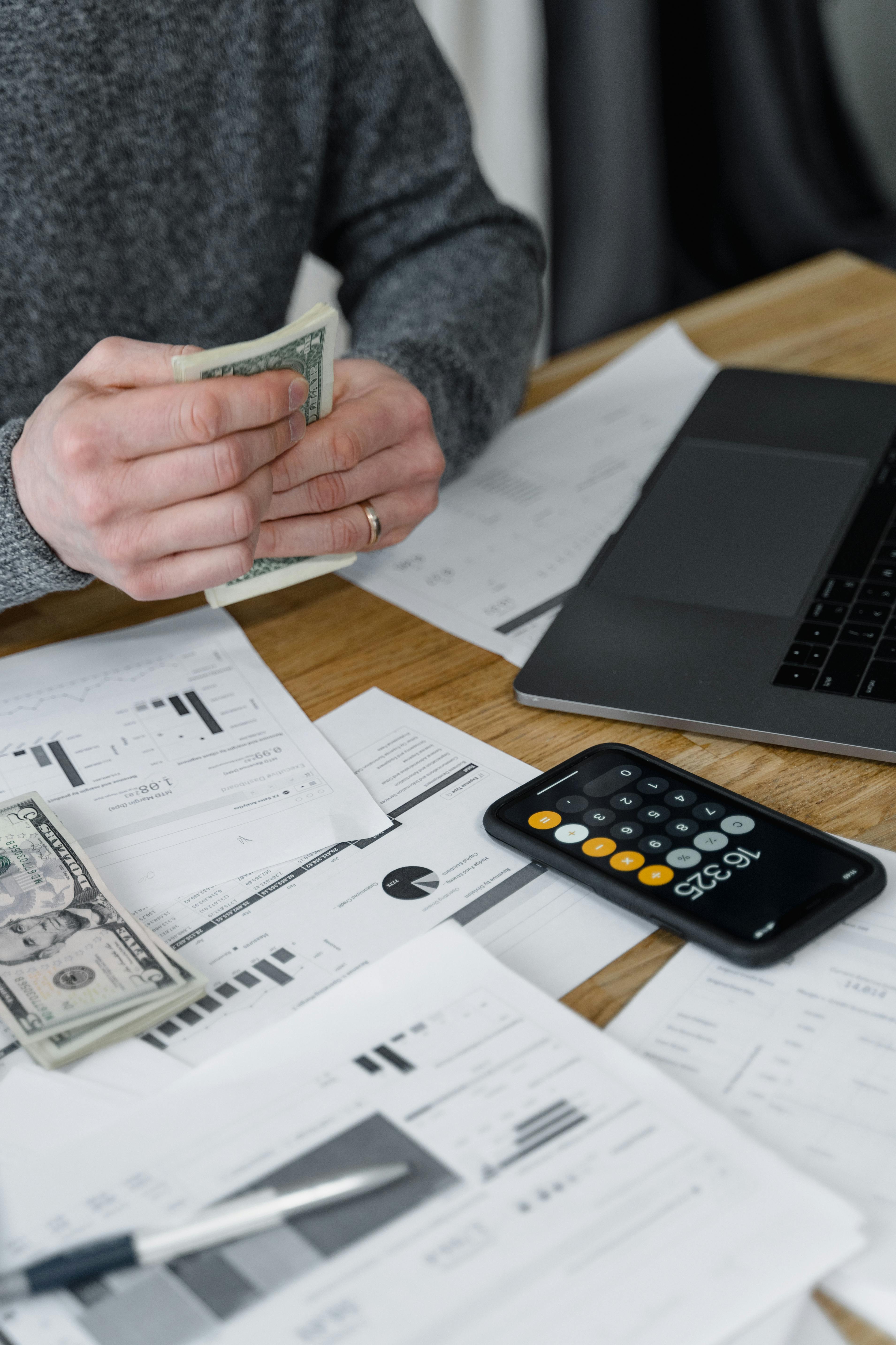 Hands counting money at a workspace with a laptop, calculator, and financial documents.